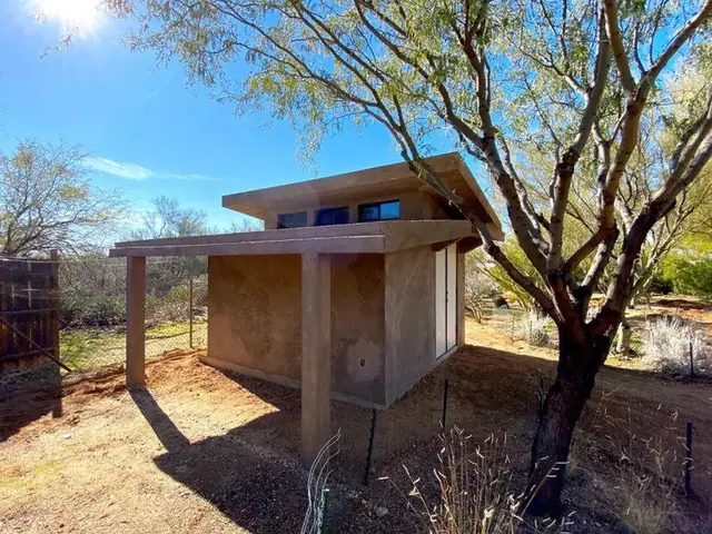 A compact completed standalone structure at studio scale, with a flat overhanging eave, one door and two windows.